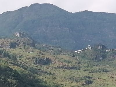 The image depicts a mountainous landscape in the city of Tả Van, Vietnam. A large mountain with dense green vegetation dominates the background. In the foreground, the terrain is characterized by rolling hills covered in grass and patches of exposed rock. Scattered throughout the hills are a few buildings, including a cluster of structures visible on a rocky peak. A statue is also seen on a similar peak. The sky is overcast.