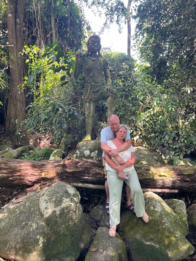 An outdoor, medium shot captures an elderly Caucasian couple posing in front of a moss-covered statue in a forest environment, possibly in Ban Bang Khao, Thailand. The man, wearing a light blue short-sleeved button-down shirt and tan shorts, stands behind the woman, embracing her from behind. He has his arms wrapped around her waist and his head leaning slightly on her shoulder. The woman, with gray hair and wearing a white sleeveless blouse and light green wide-leg pants, faces the camera with her hands clasped in front of her. She is positioned in front of the man and slightly to the right, leaning her head towards him. She is holding a mobile phone in her left hand. Both are barefoot. The statue, depicting a hunched figure with a stylized face and wearing a loincloth, is positioned behind the couple and to the left, and appears to be made of stone, covered in moss. The surroundings are lush and green with dense foliage, large trees, moss-covered rocks, and a large fallen tree trunk in the foreground. The lighting suggests it is daytime, with sunlight filtering through the leaves, creating dappled shadows. The overall scene evokes a sense of nature and ancient structures.
