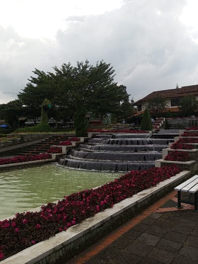 A tiered fountain flows amidst vibrant red flowers in a garden setting under a cloudy sky. The water cascades down multiple levels, leading to a pool. Lush green trees and manicured shrubbery, including a topiary peacock, complement the landscape. A building with a terracotta-tiled roof is visible in the background, adding a structural element to the scene. A white bench sits beside the lower-level flower bed.