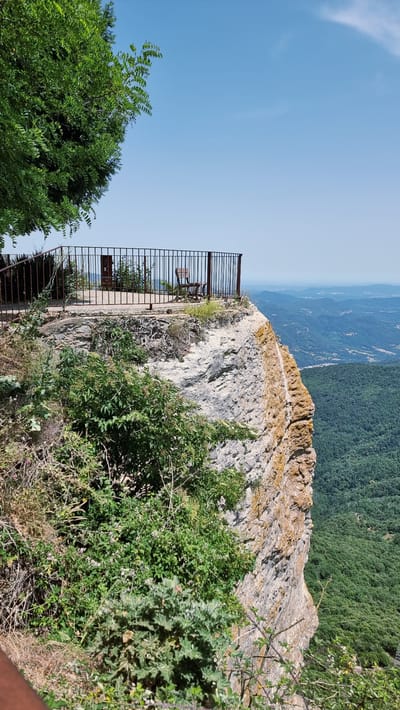 The image depicts a scenic overlook in Susqueda, Spain, with a sharp drop-off from a rocky cliff face. A metal railing encloses a paved area, where a single wooden chair and a signpost are visible. Lush green trees and vegetation frame the overlook, with a dense forest stretching out into the valley below and a hazy horizon in the distance. The sky is clear and blue, indicating a sunny day. The cliff face itself is rough and textured, showing signs of age and erosion, with patches of yellow and orange lichen adding color to the grey rock.
