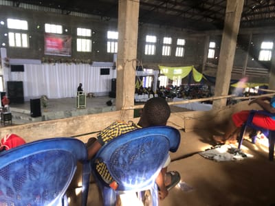The image depicts the interior of a large hall in Nteje, Nigeria, with a stage and rows of seating occupied by numerous individuals. A man speaks at a podium on the stage. In the foreground, a child wearing a checkered shirt sits in a blue plastic chair, their back to the camera. Another individual in a red shirt sits in a blue chair to the right. The hall has concrete pillars and multiple windows. Yellow fabric hangs above the seating area in the center. A white backdrop adorns the stage. A sound system is visible on the stage.