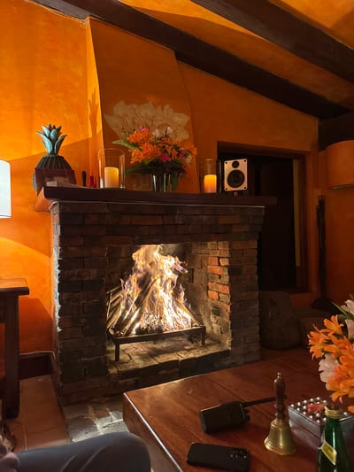 The image depicts a lit brick fireplace in a room with orange textured walls. A speaker is visible in an alcove above the fireplace. Flowers are arranged on the mantelpiece along with a candle in a glass holder. On the table next to the fireplace, there are a flower arrangement, a metal bell, a walkie-talkie, and a smartphone. A person in gray clothing is partially visible in the lower right corner. The location is in San Pablo Coapan, Mexico.