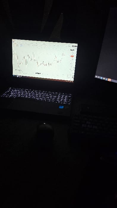 A dim, angled view shows a laptop displaying a stock chart. The keyboard is illuminated with white backlighting. A mouse sits next to the laptop, partially obscured by darkness. Another screen, possibly a monitor, is visible in the upper left corner. The surrounding environment is dark.