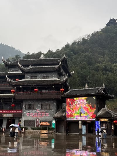The image depicts a rainy, overcast outdoor scene in Pengshui Miao and Tujia Autonomous County, China. In the foreground, a large, traditional multi-story building with dark grey brick walls, ornate dark tiled roofs with upturned eaves, and multiple balconies adorned with red lanterns dominates the left side. Adjacent to it on the right is a smaller structure featuring a prominent, brightly illuminated LED screen displaying colorful, abstract graphics, text in Chinese characters including "山海奇缘" and a time "20:00". The wet, reflective pavement of a plaza or street occupies the lower third of the frame, showing reflections of the buildings and lights.
Several individuals are present on the wet ground. On the left, a group of three to four people huddle under two open white and blue umbrellas. To the right, a solitary figure walks, partially obscured. A mobile display stand with a roasted pig is positioned in front of the main building. Various signs with Chinese characters are visible on the buildings, including a "COFFEE" sign on the rightmost structure.
In the background, a steep, densely forested mountain rises, extending upwards into an overcast sky. High on the mountain, a multi-tiered pagoda-like structure with a dark roof is faintly visible against the sky. The overall atmosphere suggests a damp, cool day.