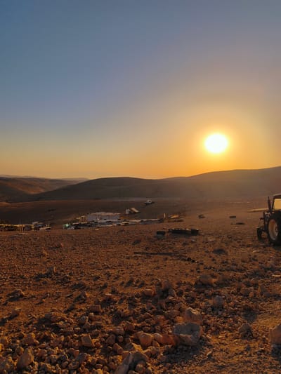 This is a vertical, eye-level, medium shot taken during sunset in the Palestinian Territories. The sun is low on the horizon, casting a warm, orange glow over the rocky, arid landscape. The foreground is dominated by numerous stones and pebbles scattered across the reddish-brown earth. In the mid-ground, a cluster of low-lying, simple structures, possibly temporary dwellings or storage sheds, are visible. A white vehicle is parked to the left of these structures, and various items, including tires, are strewn around the area. A tractor is partially visible on the right side of the image, with its rear wheel prominent. Rolling hills form the background, receding into the hazy distance under a gradient sky that transitions from a pale blue at the top to a bright orange near the horizon. The overall impression is one of a remote, undeveloped settlement in a desert environment at the end of the day. The location is identified as the city of Keidar, Palestinian Territories. There are no visible people in the image.