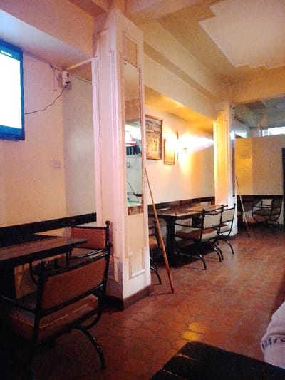 A room features tables and chairs, illuminated by a television on the left wall. Columns with mirrors stand, and framed pictures hang on the back wall. Two billiard cues lean on the columns, adding a touch of leisure. The floor is a reddish hue, and the lighting creates a warm ambiance. The scene depicts a cozy interior space.