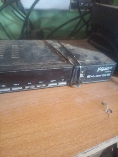 The image shows a dusty, black satellite receiver box on a wooden surface. The box has several buttons labeled "VOL-", "VOL+", "CH-", "CH+", "MENU", and "OK". On the right side of the box, there are two USB ports and a logo that appears to say "FOXCAT". The background is out of focus and appears to be a green and black patterned surface with some wires. The wooden surface in the foreground has some small holes and is textured. There are no people or animals visible in the image. The image does not contain any specific geographic markers that would allow for its location to be identified as Zot Shen, Nigeria.