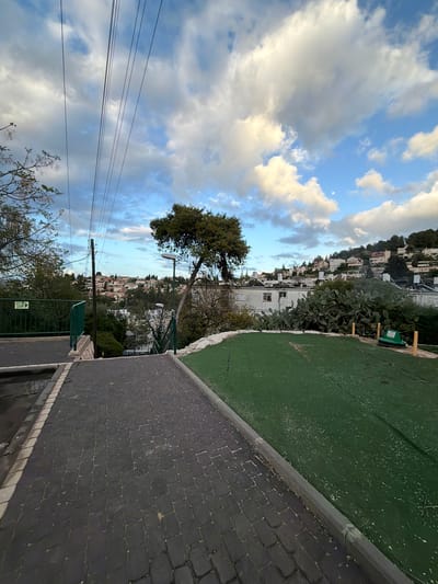 A slightly tilted view shows the Israeli city of Yokneam Illit. The foreground is occupied by a dark gray brick paved pathway. Adjacent to the pathway is a green-painted, possibly artificial, grassy area. Houses and buildings dot the hills, suggesting the city's topography. A tree is prominently positioned in the middle ground, partially obscuring the view of the town. Utility lines extend across the sky. Fluffy white clouds are scattered against a blue sky. A street light stands beside the tree, overlooking the city. A green-painted fence with a sign is visible in the lower right corner.