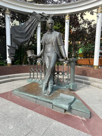 The image shows a bronze statue of a man in formal attire, holding a top hat in his left hand and resting his right arm on a decorative railing. The statue is positioned on a triangular bronze base that is set on a multi-tiered pedestal. To the left of the statue, a large abstract metal sculpture is integrated with the surrounding white, cylindrical columns and a curved white canopy. On the right side of the base, a rolled-up scroll with musical notation is depicted. The surrounding area is paved with granite tiles and red granite borders. In the background, there are trees with dark foliage, a building with yellow walls, and outdoor seating with benches. A person in a green cap is visible in the far right background. The scene appears to be an outdoor public space, possibly a park or square, in the city of Putyatino (Semivragovskaya S/A), Russia.