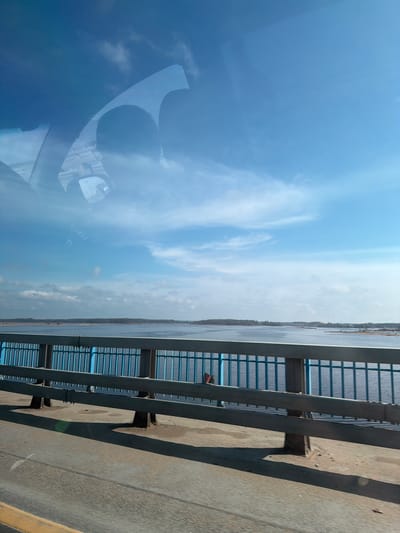 The image appears to be taken from inside a vehicle driving across a bridge. The sky and clouds are reflected in the windshield. A body of water is visible next to the bridge, with trees lining the shore. The bridge has a metal railing. The location is Bezborodovo, Russia.