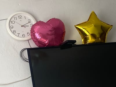 A white analog clock with black numbers and hands is mounted on a textured, light-colored wall. The clock face shows the time as approximately 3:10. To the right of the clock, a large, pink, heart-shaped balloon and a gold, star-shaped balloon are positioned on top of a black television screen. The balloons appear to be made of Mylar. A small, black object, possibly a camera or a remote control, is visible on the edge of the television screen between the two balloons. Several black wires are bundled together on the left side of the image, connected to the television or another device not fully visible. The overall environment suggests an indoor setting, likely a room in Keŋeş, Kyrgyzstan.