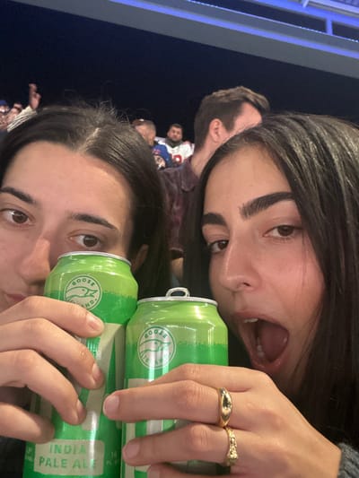 The image depicts two young adult females in the foreground, holding green "GOOSE ISLAND INDIA PALE ALE" beverage cans, situated at night within MetLife Stadium in East Rutherford, United States. The female on the left is partially visible from the nose upwards, holding one can with both hands. The female on the right is more fully visible, holding a second identical can with her right hand, her mouth open in a wide, expressive manner. She wears a plain gold-colored band and a textured gold-colored ring on her right ring finger. In the blurred background, several individuals are visible in stadium seating, including a male wearing a blue and white sports jersey. Artificial blue lighting illuminates an overhead stadium structure, contrasting with the dark night sky.