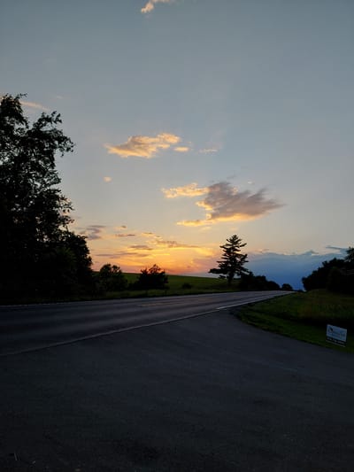 A country road curves to the right under a twilight sky. The sky is a gradient of light blue at the top, transitioning to soft orange and yellow hues near the horizon where the sun has set, casting a warm glow on scattered clouds. A silhouette of dark trees lines the left side of the road, with a few scattered trees and a lone pine tree visible in the middle distance against the colorful sky. Green fields and distant trees form the landscape. The asphalt road is dark and empty, with yellow and white lane markings visible. In the lower right corner, a small white sign with dark text is placed on the grassy shoulder. The scene appears to be in a rural area, possibly on the outskirts of Lancaster, Pennsylvania, given the presence of a local business sign with a 748 area code, which is a Pennsylvania area code. There are no people or animals visible in the image.