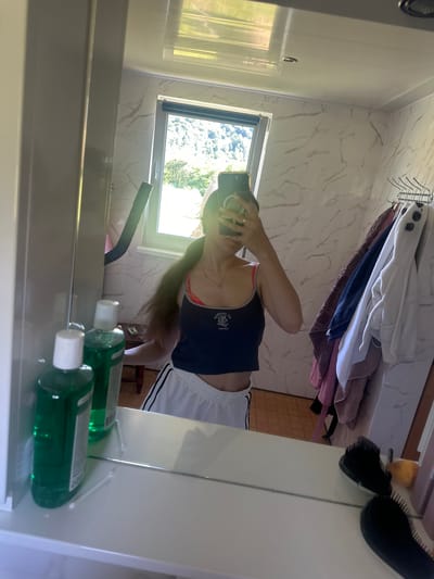 The media file shows a person taking a mirror selfie in a bathroom. The person is wearing a dark blue tank top and white shorts with black stripes. They have long brown hair that is being held back by their arm as they hold up a smartphone to take the photo. The bathroom has marble-patterned walls and a window that looks out onto a green, leafy landscape, suggesting it is daytime and possibly warm weather. On a shelf in front of the mirror are two green bottles with white caps, likely toiletries. In the reflection, there are also towels hanging on a rack and hairbrushes. The scene appears to be a domestic setting, consistent with a home bathroom. There is no visible text in the image.