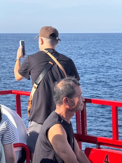 Two men are on the deck of a boat, likely in the waters off Dong Son, Vietnam. The man in the foreground is older, with short, graying hair and is looking to his right, smiling. He is wearing a black sleeveless shirt. Behind him, a younger man in a brown baseball cap and black t-shirt is holding up a smartphone to take a picture of the water. He has a bag slung across his chest. The boat has a bright red railing. The sea is a deep blue, and the sky is clear and pale blue.