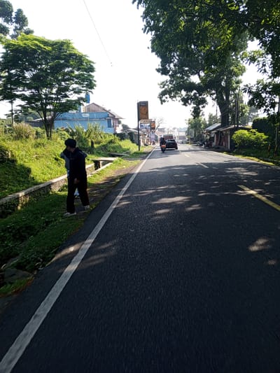 The image captures an outdoor scene on a paved road in Weddagala, Sri Lanka, under bright daylight. The perspective is from a low angle, looking down a two-lane asphalt road that recedes into the distance.
On the left side of the frame, a grassy verge borders the road, featuring a concrete drainage ditch with visible water at the bottom. Two individuals are present on this verge. The foreground figure, wearing a dark hooded top and dark trousers, stands with their back mostly to the camera, appearing to look down towards the ditch. Their left hand is raised towards their head. A second person, largely obscured behind the first and to their left, wears a lighter grey long-sleeved top. Beyond the verge, lush green vegetation, including a prominent large-leaved tree with bright green foliage, partially obscures a light blue building with a reddish-brown tiled roof. A vertical billboard-style sign, partially visible, displays the "JOTUN" logo and the word "Chaka" further down the road on the left. Overhead, thin power lines traverse the sky.
The asphalt road occupies the center and right portions of the image. A solid white line delineates the left edge of the road (shoulder), and a double yellow line is visible further down the center of the road, indicating two lanes of traffic. Dappled shadows from overhead trees are cast across the road surface, suggesting strong sunlight filtering through foliage.
In the mid-distance on the road, a dark motorcycle with a rider is visible, followed closely by a dark-colored sedan. Both vehicles are traveling away from the camera. Further down the road, additional vehicles (primarily motorcycles) and what appear to be small roadside structures are visible.
On the right side of the road, a large, dense tree with dark green foliage dominates the midground, casting significant shade. Beyond this tree, several small, single-story buildings with varied roofing (some corrugated metal) are situated alongside the road, interspersed with utility poles and overhead wires. The sky above is largely bright white, indicating overexposure in areas, but appears clear.