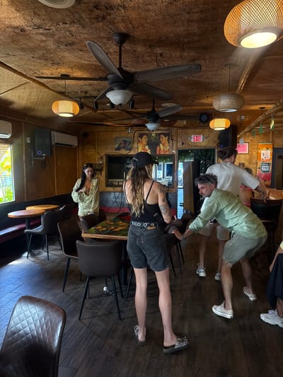 The scene depicts the interior of a casual establishment, consistent with a bar or restaurant, located in Hanalei, United States of America. The walls are covered in woven bamboo matting, and the ceiling features dark, textured natural material interspersed with bamboo matting, from which multiple dark-colored ceiling fans and woven rattan pendant lights are suspended. Two white wall-mounted air conditioning units and black speakers are present. The floor is composed of dark wood planks. Furniture includes dark brown tables, some with patterned tops, dark brown chairs with metal legs, and a built-in dark red upholstered bench along one wall. Wall decor includes framed images and an illuminated "EXIT" sign. A window on the left reveals lush green tropical foliage outdoors, while an open doorway in the background shows an exterior view.
Five individuals are visible. In the central foreground, a woman with long brown hair, wearing a black baseball cap, black tank top, dark denim shorts, and striped slide sandals, stands with her back mostly to the camera, displaying multiple tattoos on her left arm. To her right, a man with gray, curly hair and sunglasses pushed onto his head, wearing a light green button-up shirt and shorts with light slip-on shoes, leans on a chair with his right hand while holding a black object in his left. Behind him, a man with brown hair tied in a bun, wearing a white t-shirt, light shorts, and white sneakers, stands near a chair. To the left, a woman with long dark hair and sunglasses pushed onto her head, wearing a light green wrap-style top over an orange garment, stands observing the scene. A fifth person is partially visible in the far right background, wearing a pink top and white sneakers. The individuals are positioned within the space, exhibiting no clear singular coordinated action.