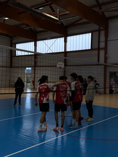 The image captures an indoor sports gymnasium in Sennecey-le-Grand, France. The central feature is a black volleyball net spanning a blue court marked with white lines. A basketball hoop with a white backboard is visible behind the net. The facility features a high ceiling with exposed wooden beams and translucent panel windows along the upper walls, allowing for natural light, supplemented by overhead artificial lighting.
On the right side of the volleyball net, five individuals are present. Four males are dressed in matching red and white short-sleeved jerseys with black shorts, displaying the text "DOLORSIT" on the shoulders and numbers such as "9" and "19" on their backs. One of these individuals has his right arm draped over the shoulder of another. A fifth male stands adjacent to this group, wearing a light-colored hooded top and dark trousers. This group appears to be in a huddle or engaged in a discussion.
On the left side of the net, further back on the court, one individual, dressed in dark attire, stands looking down at a mobile phone. Additional, indistinct figures are visible in the far background. The scene depicts activity within a sports venue.