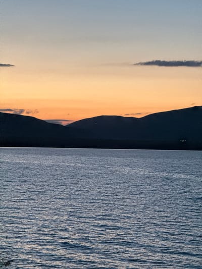 The image shows a body of water in the foreground, with gentle ripples reflecting the light. Beyond the water, a dark silhouette of mountains stretches across the horizon. The sky above is a gradient of colors, transitioning from a pale blue at the top to a warm orange and yellow hues near the mountain line, indicating either sunrise or sunset. A few wispy clouds are visible in the upper part of the sky and subtly along the mountain peaks. There are no people or specific landmarks visible that would identify the location within Tsamakaberd, Armenia. The scene is tranquil and natural.