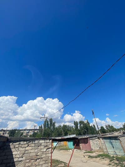 The image depicts a clear blue sky with scattered white clouds, a solitary bird in flight, and a power line stretching diagonally across the frame. Below the sky, a row of garages with metal doors of various colors is visible, situated behind a low brick wall. In the background, a multi-story apartment building and a line of tall trees can be seen. The scene appears to be an outdoor storage or parking area, likely in a residential part of Znamensk, Russia, given the architectural style of the buildings and the presence of garages. The overall impression is one of a sunny day in a suburban or urban fringe environment.