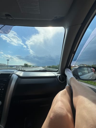 A person's legs are propped on the dashboard of a car while it is in motion on a road in Kitsjanovo, Russia. The sunny sky and the car's dashboard and interior are visible.