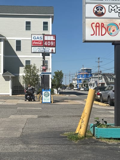 A man on a motorcycle is parked at a gas station in Hampton, United States of America. The gas price sign displays "GAS Coca-Cola 409". Another sign above it reads "PATRIOTS CORNER". To the right, a portion of a sign with images of a panda and a muscular man is visible. A bright blue building under construction is in the background, along with other houses and power lines. A silver car is parked on the side of the road. The ground is paved asphalt with some cracks and patches. A bright yellow pole and a turquoise planter with green plants are in the foreground. The sky is clear and blue.