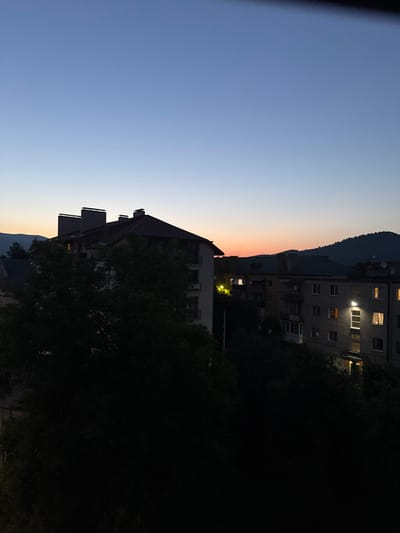 The image shows an outdoor scene at dusk or dawn in Svaliava, Ukraine. The sky transitions from a deep blue at the top to a soft orange and pink hue near the horizon, indicative of twilight. Silhouetted against the sky are rolling hills covered with trees.

In the foreground and midground, several multi-story apartment buildings are visible. Some of these buildings are partially obscured by dense, dark green foliage, likely trees. The buildings themselves are utilitarian in design, with visible windows and balconies. A few windows are illuminated, suggesting that people are inside their apartments, with lights on. One building on the right has exterior lighting illuminating a section of its facade. Another building on the left, partially obscured by trees, has an upper section with visible chimneys.

The overall impression is of a residential area within a town or city situated in a hilly or mountainous region, observed during the transitional light of twilight. There are no people or active interactions visible in the image. The scene is static and captures the quietude of the hour.