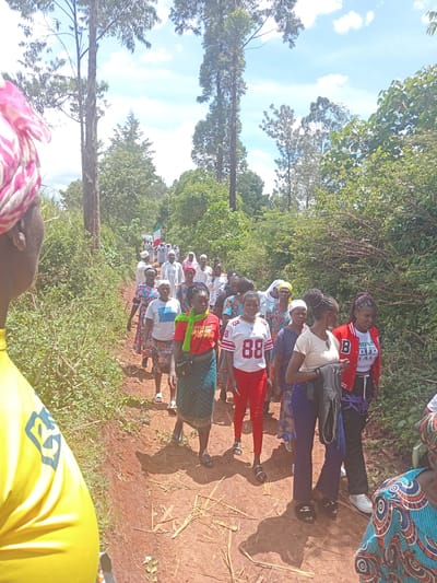 The image displays a daytime scene in Butali Chegulo ward, Kenya, featuring a procession of approximately 25-30 individuals, predominantly women, walking along a reddish-brown dirt path. The perspective is from behind and slightly to the right of an individual wearing a yellow garment and a pink and white patterned headwrap.

The environment is rural, characterized by dense green vegetation flanking both sides of the path, including bushes, shrubs, and tall, slender trees with sparse foliage. The sky overhead is bright blue with scattered white clouds.

The individuals in the procession are walking in a loose single file or small clusters. Towards the rear of the visible group (furthest from the camera), several individuals wear long white robes, some with head coverings. One person in white carries a flag with vertical green, white, and red stripes. Closer to the camera, the attire is varied and casual, including patterned dresses, skirts, t-shirts, and long pants. Specific examples include a woman in a red and white jersey with the number '88' on the front, another in a red varsity-style jacket, and a woman in a red t-shirt with a green scarf. Footwear observed includes sandals and sneakers. Individuals appear to be moving forward, with some looking ahead and others glancing to the side. Shadows indicate the sun is overhead or slightly angled.