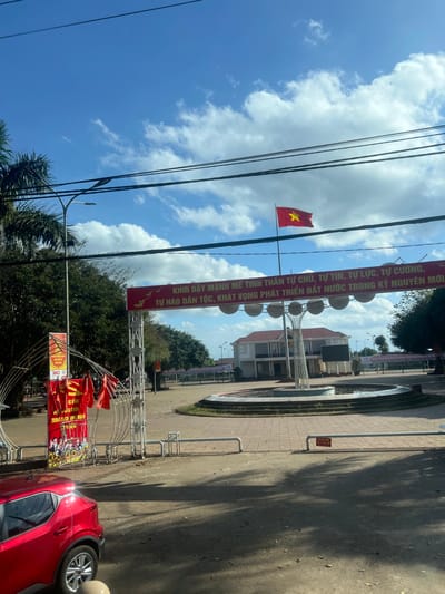 The image depicts a public square or town center in Trung Hồ, Vietnam, under a partially cloudy blue sky. In the foreground on the left, a red Nissan Kicks SUV is partially visible. The midground features a white metal archway entrance adorned with multiple small red Vietnamese flags. A prominent red banner with yellow Vietnamese text and illustrations of doves and gears is stretched horizontally across the archway, reading "STRONGLY AROUSE THE SPIRIT OF SELF-MASTERY, SELF-CONFIDENCE, SELF-RELIANCE, SELF-RESILIENCE, NATIONAL PRIDE, ASPIRATION TO DEVELOP THE COUNTRY IN THE NEW ERA." To the right, a large circular dry fountain or planter contains a modern, multi-branched sculpture with spherical elements. A "No Parking" sign in Vietnamese is visible on the paved ground near the fountain. In the background, a two-story building with a red-tiled roof and white walls stands at the far end of the square. To the right of this building, a tall flagpole displays a Vietnamese national flag (red field with a yellow star). The square appears empty of people. Palm trees and other foliage are visible on the left and in the distant background. Multiple dark overhead wires cross the upper portion of the frame.