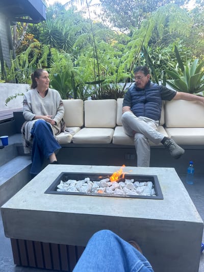 A man and woman are sitting on an outdoor sectional couch. The man is wearing a blue puffy jacket and light-colored pants and is reclining back. The woman is wearing a gray sweater and jeans. A fire pit is in front of them, filled with white rocks and burning with an orange flame. A water bottle sits on the ledge next to the fire pit. Lush green foliage and plants surround the seating area.