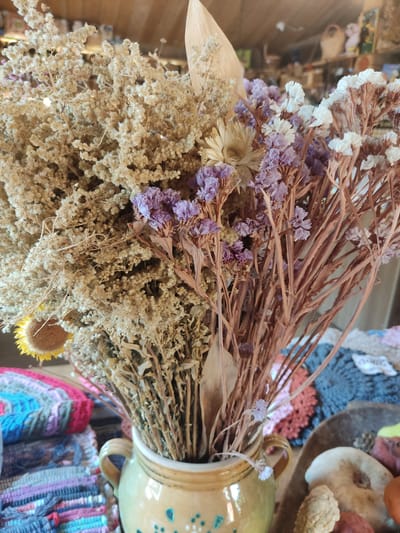 A rustic bouquet of dried flowers and plants is centrally depicted in a close-up shot, situated in Staraya Russa, Russia. The bouquet primarily features voluminous clusters of light brown, fluffy dried foliage, interspersed with slender stems bearing small purple and white dried flowers, resembling statice. A single dried yellow sunflower head with a brown center is visible on the lower left side of the arrangement. The flowers are contained within a light yellow-brown ceramic vase with two small handles, decorated with simple blue-green painted motifs near its base. To the left of the vase, a stack of multi-colored, woven or crocheted textile items, displaying horizontal stripes in shades of pink, blue, and grey, occupies the foreground. To the right of the vase, a shallow wooden bowl contains various natural decorative elements, including a white, irregularly shaped object, a reddish spherical object, and smaller, textured items. The background is softly blurred, revealing the interior of what appears to be a shop or market stall with wooden shelving and other artisanal products. No individuals are present in the frame.