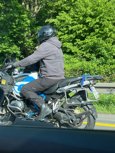 The image shows a motorcyclist riding on a road, with dense green foliage visible in the background. The motorcyclist is wearing a dark grey hooded sweatshirt with the word "Deus" printed on the back, black pants, and a dark helmet. The motorcycle is a BMW R1250GS with blue and white accents. The license plate is partially visible, with the letters "ST-KD" visible. The location is Sankt Augustin, Germany. The rider appears to be maneuvering the motorcycle on the road. A yellow line is visible on the edge of the road. The photograph appears to have been taken from inside a vehicle.