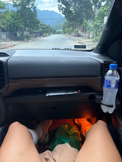 The image shows the interior of a car, focusing on the dashboard and passenger area. A bottle of "TAI CHE TOI" Coca-Cola water sits in a holder on the dashboard. A document is tucked into a slot in the dashboard. The passenger-side footwell contains a green plastic bag, a pair of shoes, and bare legs. The windshield reveals a view of Minh Tân Commune, Vietnam, with trees, buildings, and mountains visible.