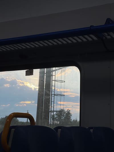 The image shows a view from inside a train compartment, looking out the window. Outside the window, there are clouds illuminated by the setting sun, and a tall, industrial-looking structure, possibly a power line pylon or antenna tower, is visible. Some trees and power lines can also be seen in the distance. Inside the train, the dark blue seats and a yellow handrail are in the foreground. A small sticker with a QR code is affixed to the window. The lighting suggests it is either early morning or late evening. The location is stated to be Pereyaslavka, Russia.