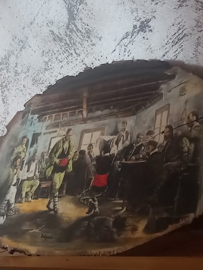 The image depicts a painting on a wooden surface, likely a slice of a tree trunk, with a rough, textured edge along the top. The painting portrays an indoor scene with multiple figures. In the foreground, a man in a green jacket, black vest, and red sash is centrally positioned, facing towards the viewer and gesturing with his hands. He appears to be in motion, possibly dancing or demonstrating something.
Behind him, several other men are gathered. Some are seated around a table in the background, engaged in conversation or observation. Others are standing. The room has a rustic interior with wooden beams supporting the ceiling and windows visible on the wall. The overall atmosphere seems to be one of a social gathering or a traditional event.
In the lower-left corner of the painting, the word "Bulgaria" is inscribed. The painting is set against a textured, light-colored background, which appears to be the natural surface of the wood. The foreground also shows the wooden surface. There is no specific indication of the city of Radishevo in the visual details of the painting itself.