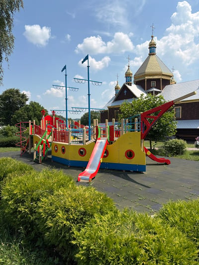 The image shows a colorful children's playground designed as a pirate ship. The ship structure is primarily yellow and blue with red accents, featuring multiple levels, slides, a spiral staircase, and a crane-like element. The playground is situated on a black rubberized surface with green grassy areas and manicured bushes bordering it. In the background, a large, ornate church with golden domes and crosses is visible. The sky is blue with scattered white clouds. The location is identified as Triitsia, Ukraine. There are no people visible in the image.