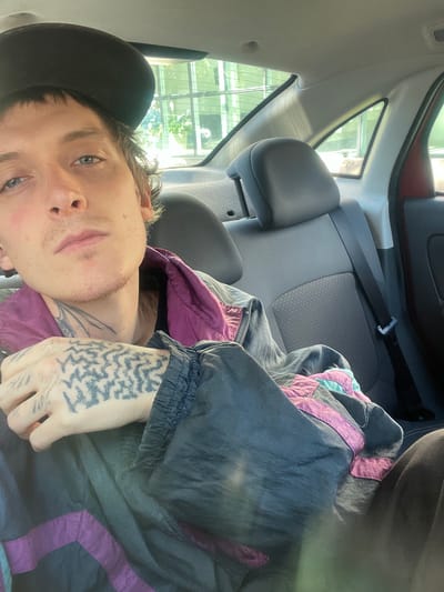 A medium shot depicts a person in the back seat of a car. The individual is lying down, wearing a black baseball cap and a jacket with purple and teal accents. Visible on the person's hand is a tattoo. The car's interior, including the headrest and seatbelt, are visible. A window shows a view of what seems to be a building or complex of buildings with vertical structures. The location is not specifically discernable as Olivos, Argentina, based on the image.