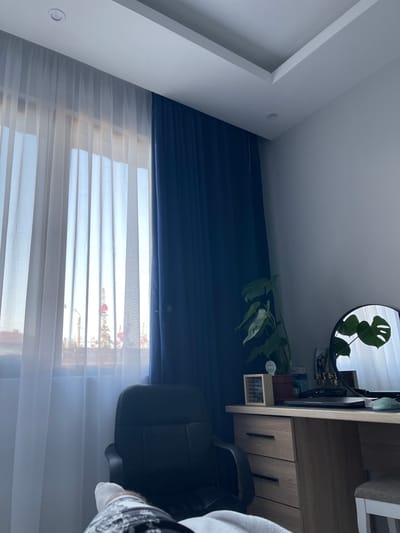 The image depicts an indoor scene, likely a home office, in Vedrare, Bulgaria. A desk with a wooden finish and drawers is visible on the right side of the frame. A black office chair is positioned in front of the desk. A large dark blue curtain covers the upper portion of a window, while sheer white curtains are draped below, allowing natural light to filter through. On the desk, a round mirror reflects a Monstera plant with distinctively shaped leaves. Various small objects are placed on the desk and on a shelf above it, including what appears to be a framed piece of paper or a small calendar with text, and decorative items. The overall lighting suggests it is daytime, with the light coming through the window. The scene is static, with no activity explicitly taking place. The room appears tidy and organized.