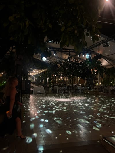 The image depicts a dimly lit outdoor event space in Beit Hanan, Israel. A woman in a black dress and high heels stands on the left, holding a drink. The ground is tiled and illuminated by scattered light patterns, likely from disco balls or similar lighting effects. In the background, tables are set for a formal event, with white tablecloths and chairs. String lights are visible in the mid-ground, and large trees with foliage cast shadows. The overall atmosphere suggests a nighttime gathering or party.