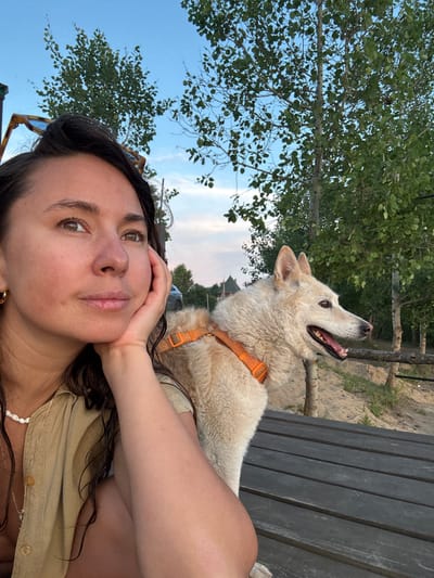 A medium shot captures a woman and a dog sitting on a wooden bench outdoors. The woman is in the foreground on the left, leaning her head on her hand and looking to the left. She has long, dark, wavy hair and is wearing a tan button-up shirt and a white beaded necklace. Her right arm is resting on the dog. The dog, a light-colored husky mix, is sitting next to her, facing to the right with its mouth open. It is wearing an orange harness. The background shows trees and a sandy area, consistent with a park or natural setting. The sky is blue with some clouds. The image appears to have been taken in the city of Kamysh, Russia.