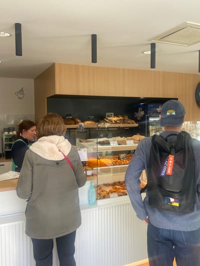 The scene depicts the interior of a bakery or patisserie located in Montigny-le-Bretonneux, France. Three individuals are present: one female employee behind a light wood counter, a female customer facing the counter, and a male customer positioned to the right of the female customer, also facing the counter with his back to the camera.
The female employee, with reddish-brown hair, wears a dark top and a light green apron, engaging with the female customer. The female customer wears a khaki-colored long jacket with a cream-colored fleece lining visible at the collar and a red strap across her shoulder, over dark pants. She stands directly in front of the counter.
To the right, the male customer wears a grey long-sleeved shirt, dark blue jeans, and a navy blue baseball cap with a yellow and green multi-elephant logo on the front. He carries a black backpack with two keychains attached; one is blue with white text "POUR ESTACA" and red airplane graphics, the other is red with white text "REMOVE BEFORE FLIGHT".
Behind the counter and customers, glass display cases showcase various baked goods, including breads, pastries, and what appear to be individual desserts or tarts. A blue bottle of hand sanitizer is visible on the counter. Above the display cases, shelves hold additional bread items, and a refrigerator with an "Orangina" logo is visible. The walls are light wood paneling above dark shelving, with a stylized coffee cup silhouette decal on the left wall. The ceiling is white, featuring multiple dark cylindrical track lights and a rectangular air conditioning vent.