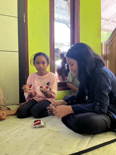 In a brightly lit room within Desa Batu Layar, Indonesia, three individuals are seated on a light-colored, quilted mattress playing UNO. The room features a vibrant lime green wall adjacent to a dark brown door frame and a window. Through the window, an outdoor scene with a corrugated roof and foliage is visible, along with reflections of other individuals.
On the left, a young female with dark hair pulled back, wearing a light pink t-shirt with a black graphic and dark pants, sits cross-legged. She holds a fan of UNO cards in her left hand and a smaller stack in her right, directly facing the viewer with a neutral expression. She wears a light-colored necklace and bracelets on her right wrist.
To her right, partially obscured by the frame, another individual's arm is visible, wearing a beige sleeve.
On the right side of the image, a second young female with long, dark hair, wearing a dark blue long-sleeved top and dark pants, sits cross-legged. She is looking down and to her left, focused on the cards in her hands or on the mattress, with a slight smile. Her bare feet are visible.
Between the two main subjects, a stack of UNO cards, face down, rests on the mattress. The overall scene depicts a casual game in an indoor setting.