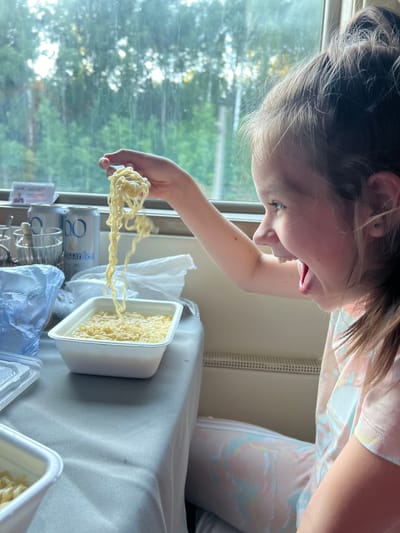A young girl with brown hair pulled back in a ponytail, is sitting in front of a window and appears to be eating noodles. She is holding a fork full of noodles up to her open mouth and her eyes are wide with excitement. The window behind her shows a blurry green forest. On the table in front of her are two white containers of noodles, a plastic bag, and two cans of what appear to be beer. The girl is wearing a pink t-shirt with a colorful swirled pattern. The scene appears to be in a train car, given the window view and the presence of a radiator below the window. The specific location in Kizner, Russia, cannot be determined from the image alone.