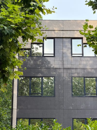 This is a low-angle, exterior shot of a modern building facade partially obscured by lush green foliage. The building is clad in a combination of light-colored, textured stone and dark, textured tiles arranged in a grid pattern. Multiple windows with dark frames punctuate the facade, reflecting the surrounding trees and sky. Some windows appear to be large, rectangular panes, while others are smaller and square.
The foliage, primarily large-leaved trees, dominates the foreground and edges of the frame, creating a sense of being enveloped by nature. Sunlight dapples through the leaves, casting highlights and shadows on the building and the leaves themselves. The sky visible in the upper left of the frame is a pale, clear blue, suggesting a bright, likely sunny day. The overall mood is serene and organic, with the man-made structure juxtaposed against the natural elements. No people are visible, and there is no visible text. The location is identified as Maisons-Alfort, France.