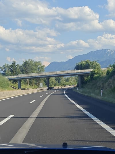 The image depicts a highway scene on a sunny day with scattered clouds. A highway overpass with a pedestrian walkway stretches across the road, flanked by trees and vegetation. In the distance, a mountain range is visible. The perspective is from a vehicle traveling on the highway, with the road markings and the front of the vehicle visible in the foreground. The scene suggests a rural or semi-rural setting. The location is identified as Schlierbach, Austria.