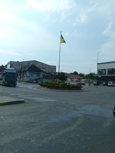 This is a street-level, medium-distance photograph of a roundabout in Tiachiv, Ukraine. The sky is partly cloudy and appears to be daylight, possibly late morning or early afternoon. The scene is a busy intersection with several vehicles.
On the left, a black Volvo semi-truck with "Lysa-trans" written on its side is moving through the roundabout. In the center of the roundabout, a patch of green bushes is surrounded by asphalt. A Ukrainian flag with a trident emblem flies on a flagpole in the background.
Several buildings line the streets around the roundabout. On the far right, a modern building with "CIAO!" and "FIMKA" signage is visible. To its left, a street sign points towards "UZHHOROD" and "TRAIN STATION" (ВОКЗАЛИ in Ukrainian). A white van is parked in front of the modern building.
Other vehicles, including red and black cars, are also navigating the roundabout. The asphalt surface of the road shows some wet patches, suggesting recent rain. The overall impression is a typical day at a provincial intersection in Ukraine. The visible text indicates commercial activity and directions to nearby locations.