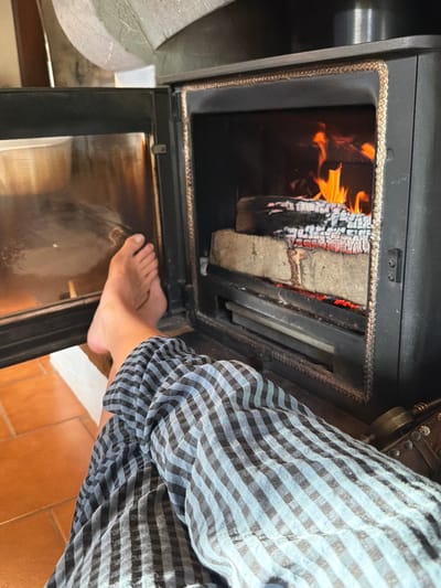 An indoor scene within a structure in Roquiague, France, depicts a dark-colored wood-burning fireplace and the lower extremities of an individual. The fireplace's open firebox contains actively burning logs, displaying orange flames and red embers over a metal grate. A light-colored seal outlines the firebox opening. To the left, a glass door exhibits reflections from the immediate environment. The bare feet of one individual are positioned on a white rectangular surface directly in front of the fireplace, indicating proximity to the heat source. The individual wears blue and black checkered trousers. The floor is composed of reddish-brown terracotta tiles. A dark, studded object is partially visible in the lower right foreground.