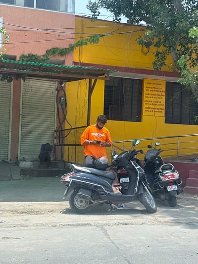 A man in an orange t-shirt stands holding a smartphone in front of a yellow building in Ajmer, India. The man is looking down at his phone. He is wearing dark pants. In the foreground, two motorbikes are parked. One is dark blue and the other is black. A person in a blue shirt is sitting on the steps of the building to the left. The building has a pink facade on the left side and a yellow facade on the right side. There is a green awning over the entrance. A sign with Hindi text is posted on the yellow wall. The street in front of the building is paved.