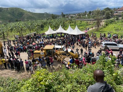 The image depicts a large gathering of people in Sake, Democratic Republic of Congo, at the coordinates -1.571, 29.046. The scene is situated on a dirt road in a rural area, with a dense banana plantation to the left. The gathering is arranged around two large yellow Caterpillar construction vehicles, one of which is a grader. There are three white tents to the right of the grader, and a crowd of people surrounding them. Several motorcycles are parked around the crowd, along with two white SUVs. The individuals in the scene are predominantly male, wearing casual attire. They appear to be observing the construction vehicles, potentially during an event or ceremony. Several individuals stand by the edge of the crowd, seeming to be in conversation with each other. The image suggests an event related to construction or infrastructure development in the region. The green vegetation and dirt road in the image further emphasize the rural environment.