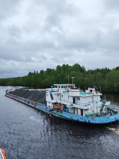 A long, white and blue barge filled with dark material, possibly coal, navigates a wide river in Depo, Russia. The barge is multi-level with multiple windows, life preservers, and a Russian flag. Green trees line both sides of the river under an overcast sky. The water is dark with visible ripples created by the barge's movement.